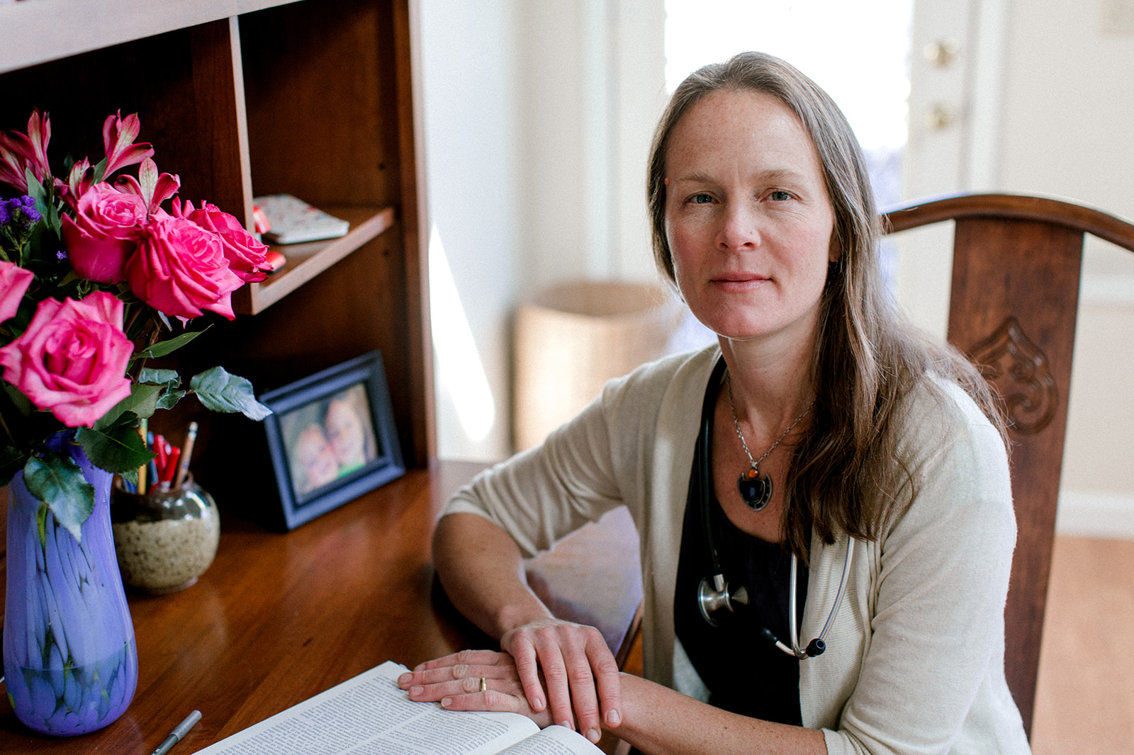 doctor-ingrid-bauer-of-five-flavors-herbs-sitting-at-desk-with-book-and-vase-of-flowers-looking-at-camera