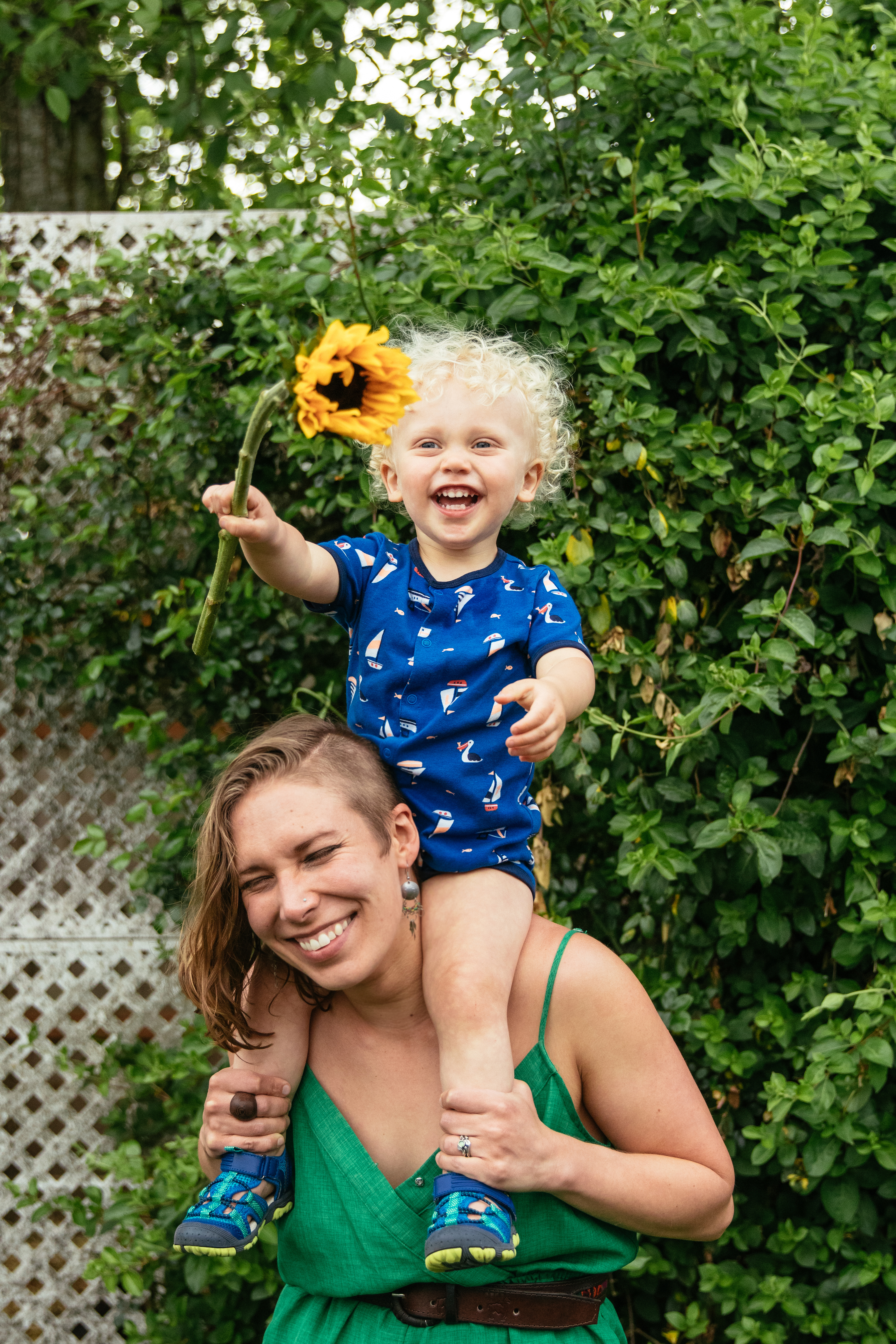 woman-in-green-jumper-holding-blonde-curly-haired-toddler-on-shoulders-holding-sunflower