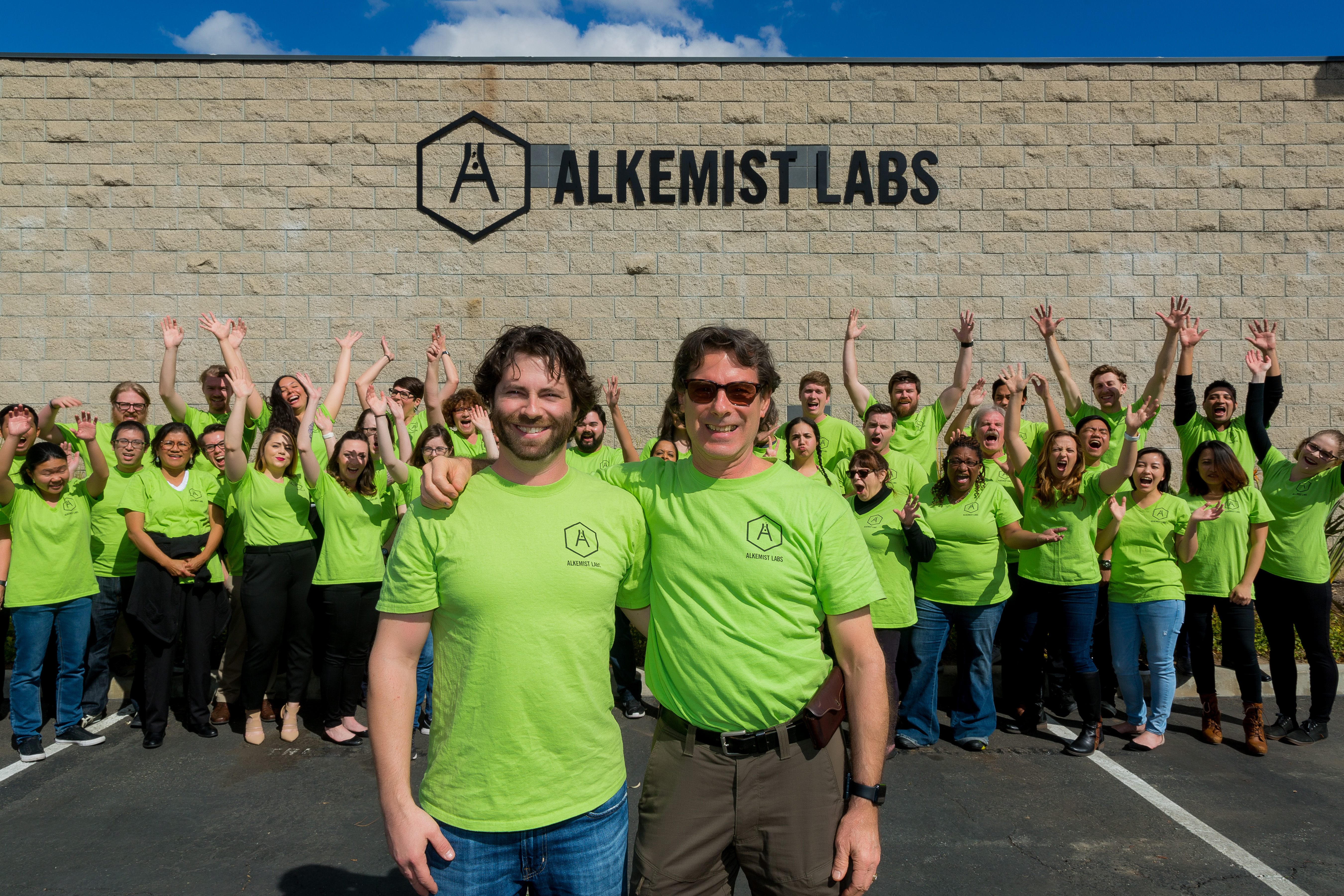 alkemist-labs-staff-photo-with-about-employees-standing-in-front-brick-wall-with-lab-name-and-logo-with-co-founders-standing-in-front