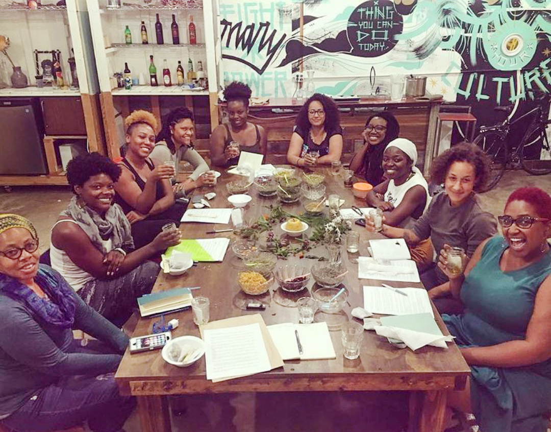 Group of Black women sitting around table at Black Power herbalism workshop with notes, bowls of herbs, and beverages, smiling at camera.
