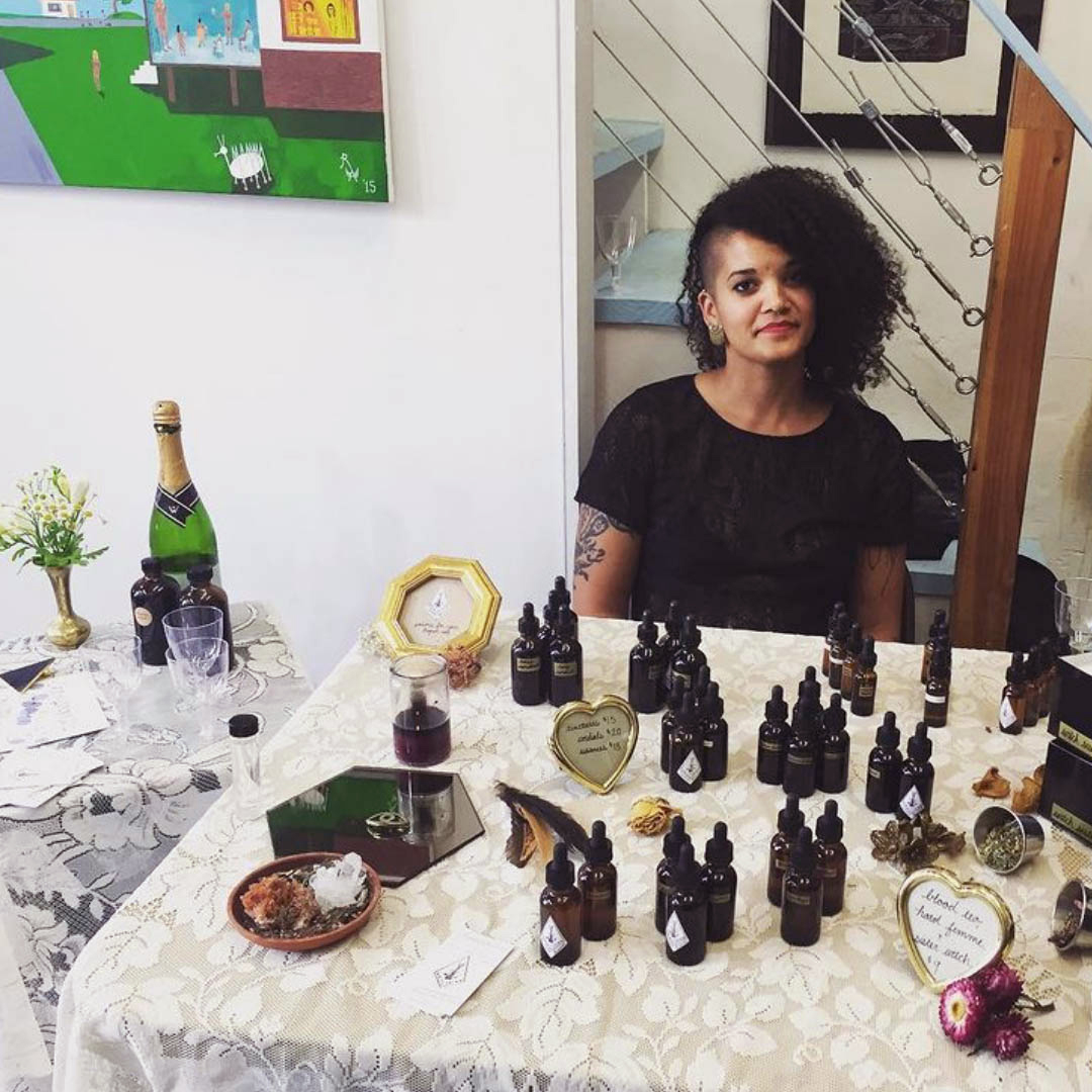 Meghan Elizabeth, young Black woman with natural hair sitting at table at maker fair with amber bottles of homemade tincture bottles on table.