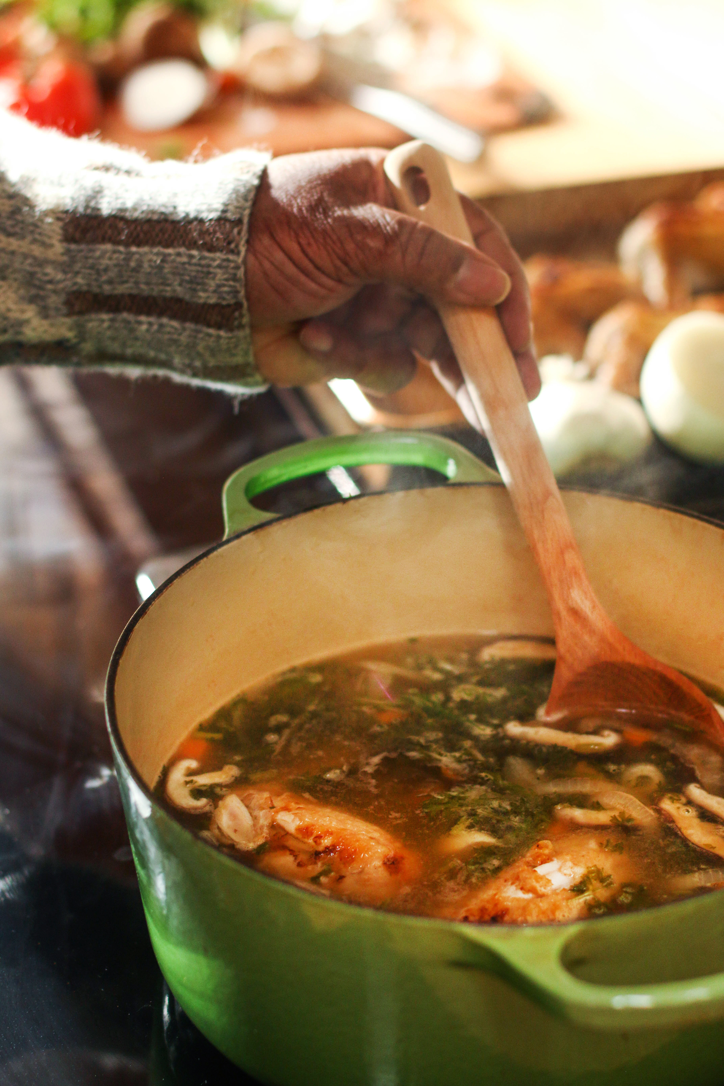 black-person-wearing-cozy-sweater-standing-over-stove-stirring-soup-with-herbs-mushrooms-vegetables-in-green-enamel-dutch-oven-using-wooden-spoon