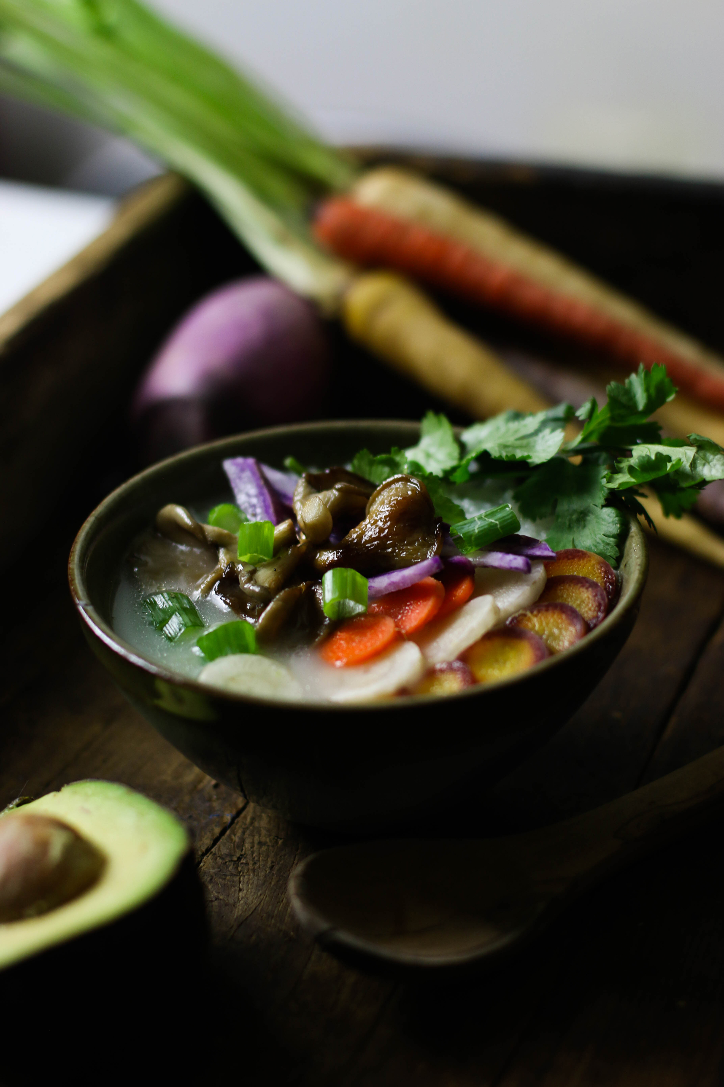 Bowl of congee with vegetables and herbs on top surrounded by an avocado and carrots