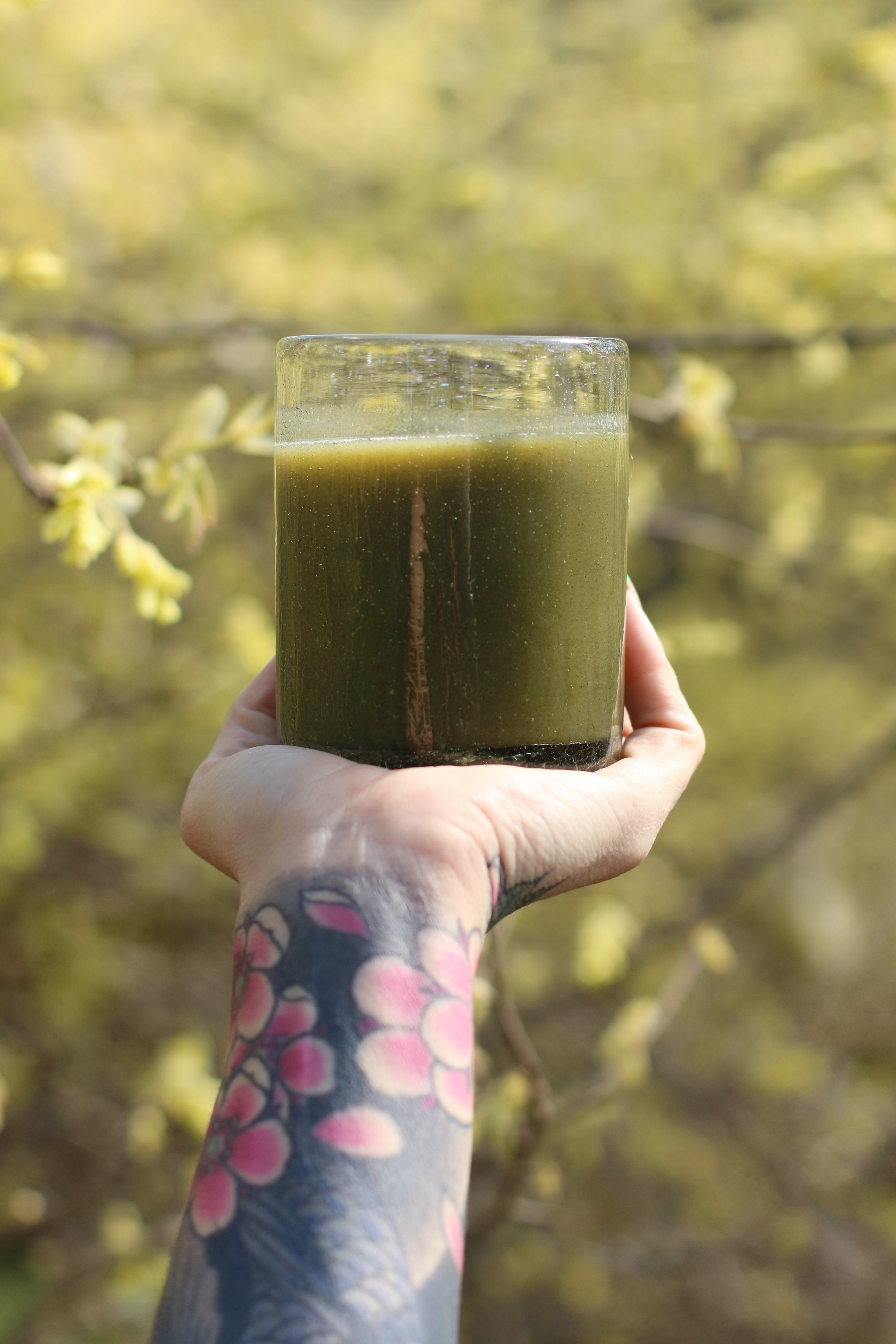 tattooed-arm-extended-palm-up-holding-glass-of-green-juice-with-trees-in-background