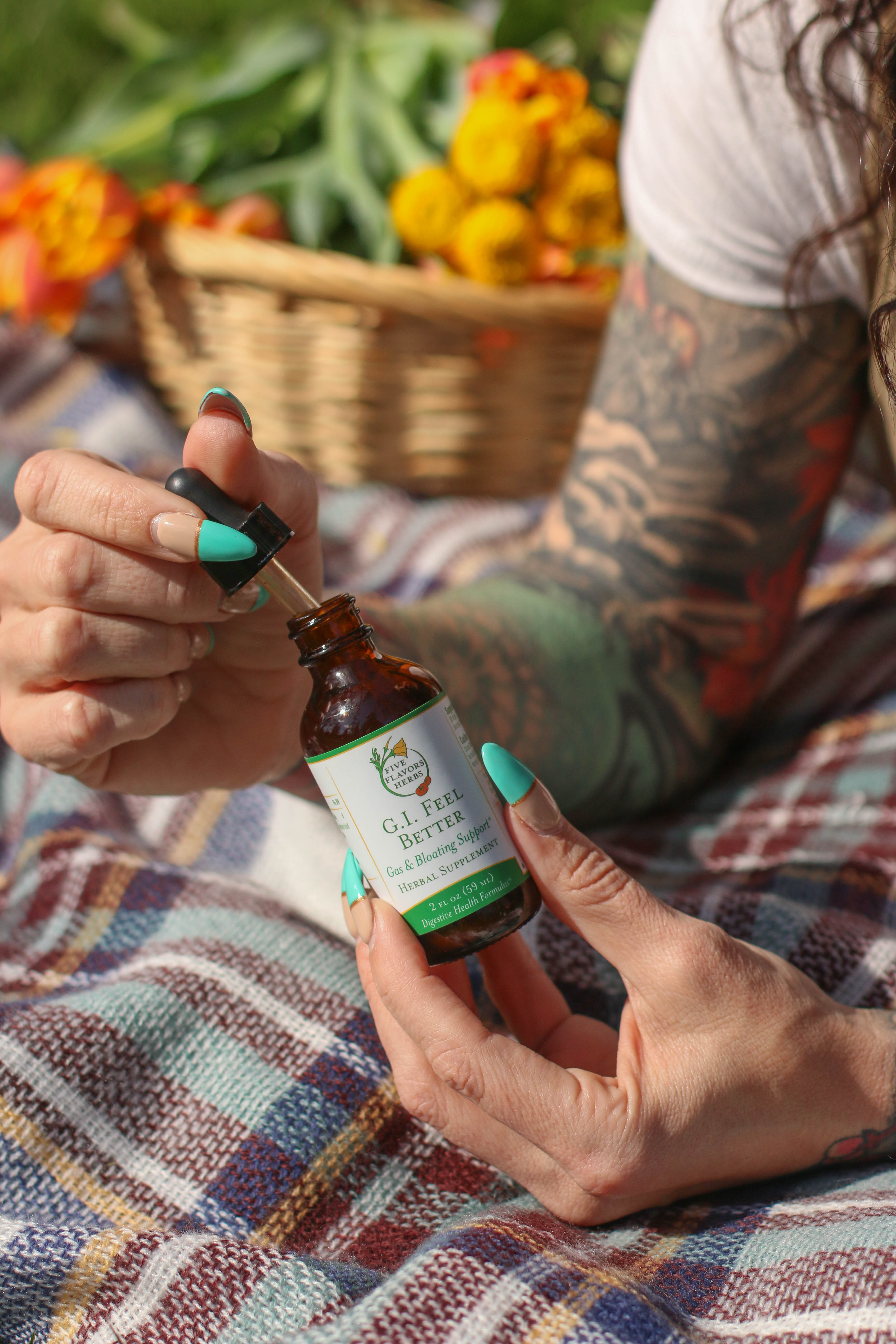 woman-with-long-painted-finger-nails-and-arm-sleeves-of-floral-tattoos-sitting-outside-on-picnic-blanket-holding-open-dropper-bottle-of-gi-feel-better-tincture-for-liver-health-and-digestion