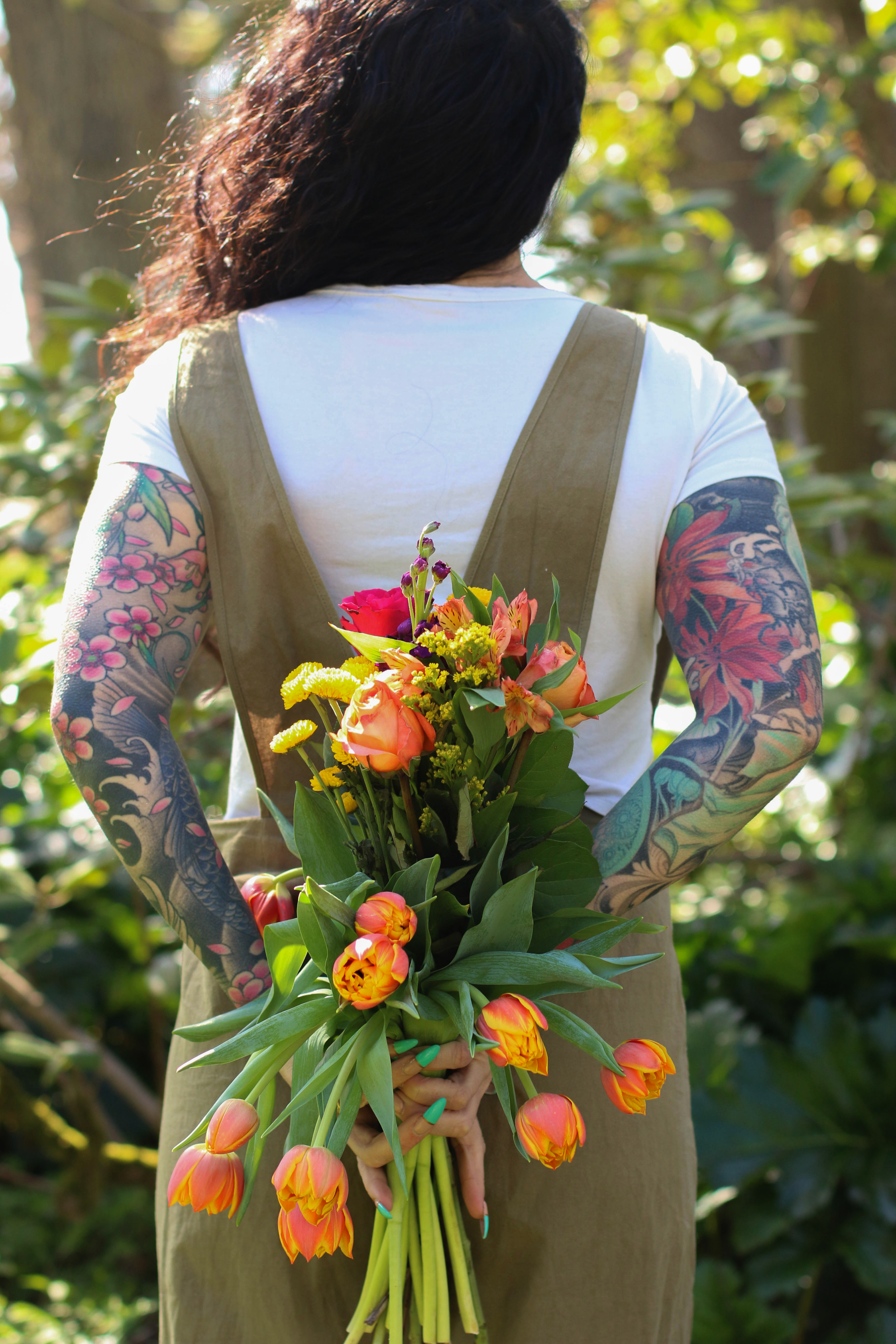 young-woman-with-tattooed-arm-sleeves-standing-outside-facing-away-from-camera-holding-big-bouquet-of-fresh-spring-flowers-behind-her-back