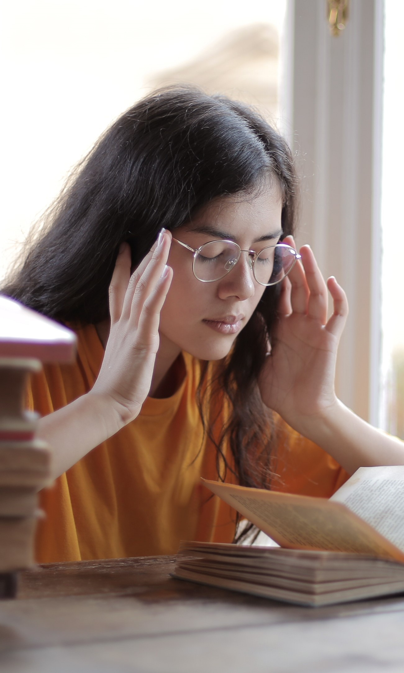 young-woman-wearing-glasses-surrounded-by-books-with-eyes-closed-and-hands-on-her-temples-appearing-to-have-a-headache