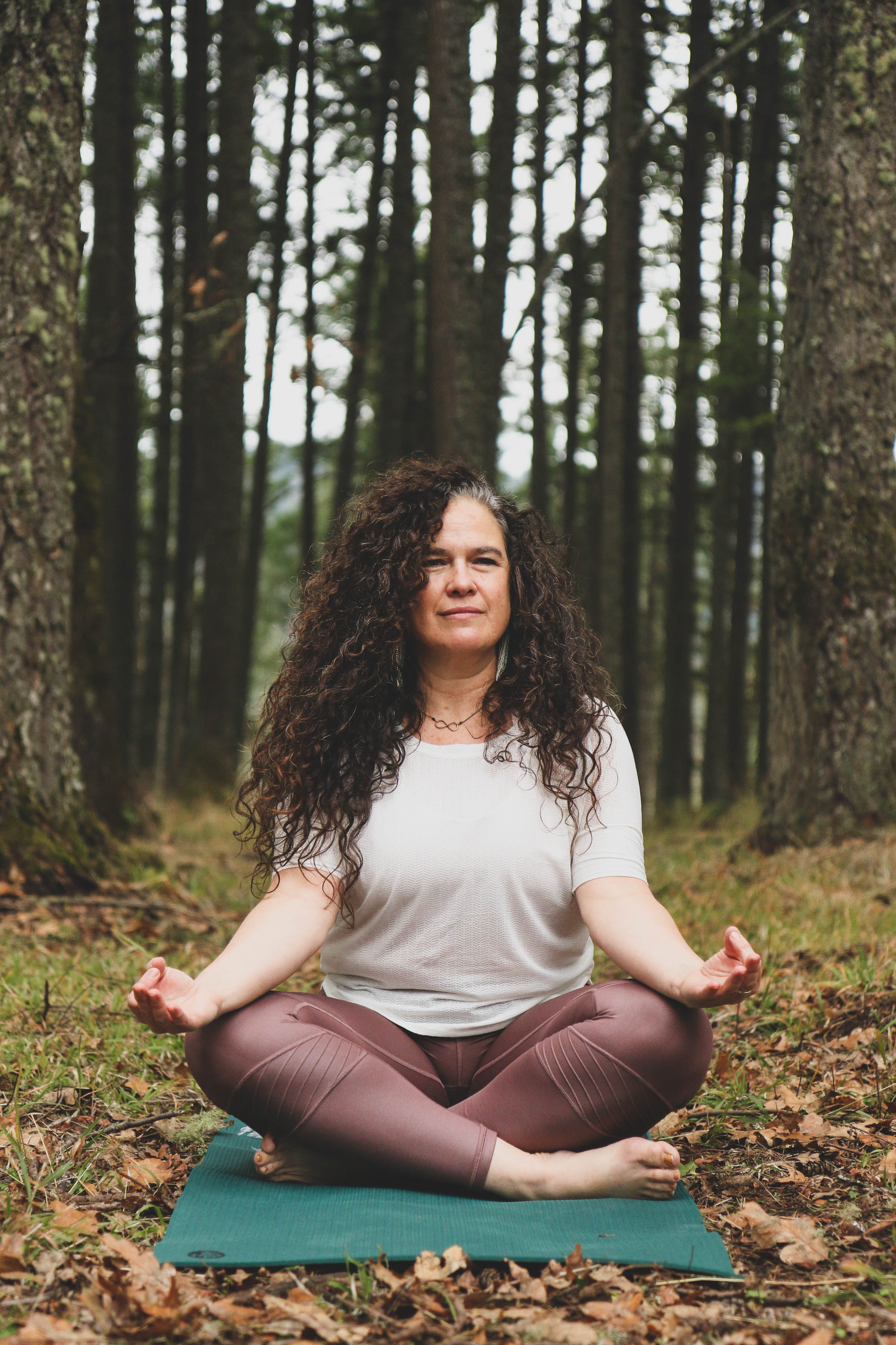 middle-aged-woman-with-long-dark-curly-hair-with-grey-streaks-wearing-white-top-and-mauve-yoga-pants-sitting-in-lotus-pose-on-yoga-mat-in-autumn-pine-forest