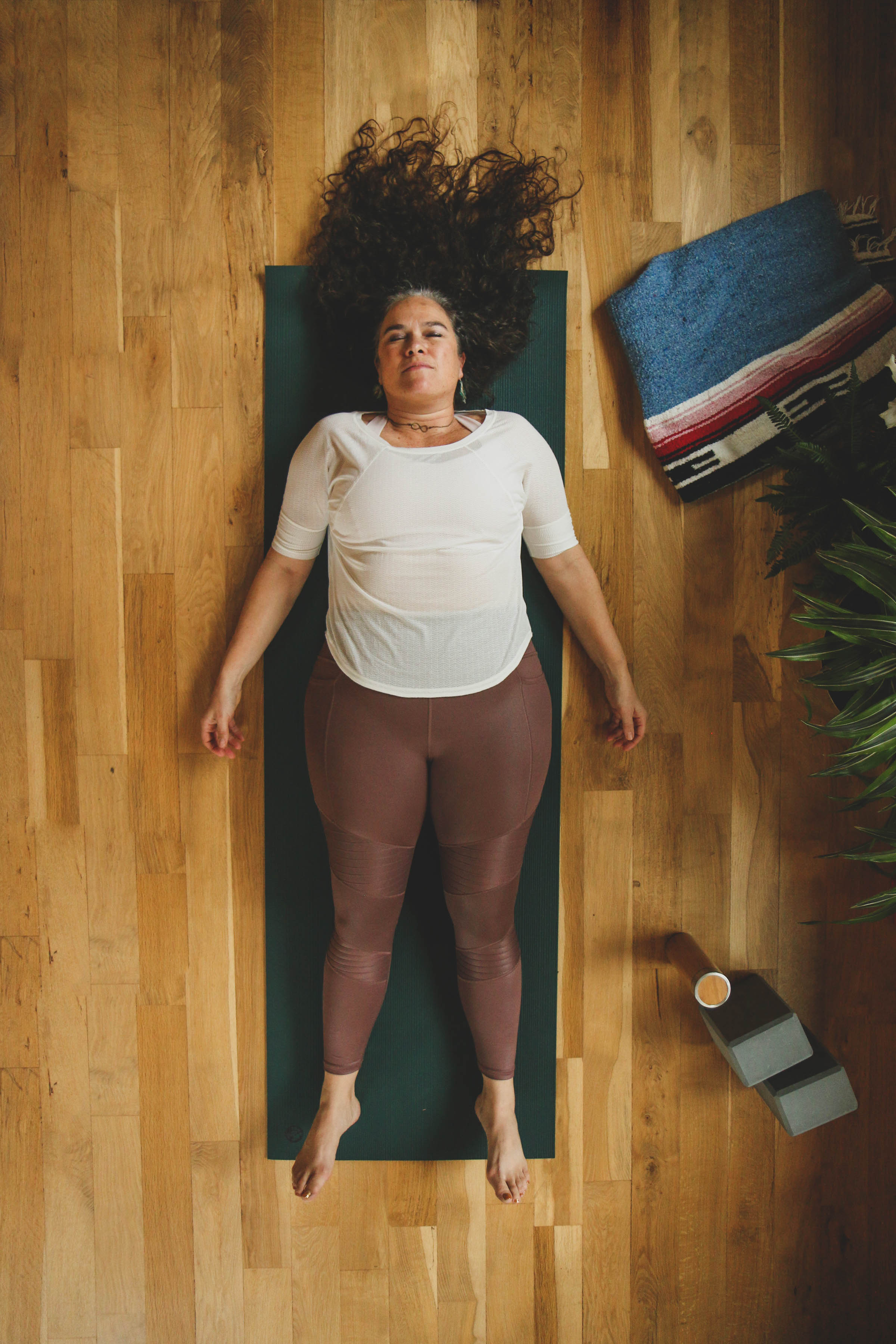 women-with-long-dark-graying-hair-lying-on-back-on-yoga-mat-on-wood-floor-with-plants-and-yoga-props-to-side