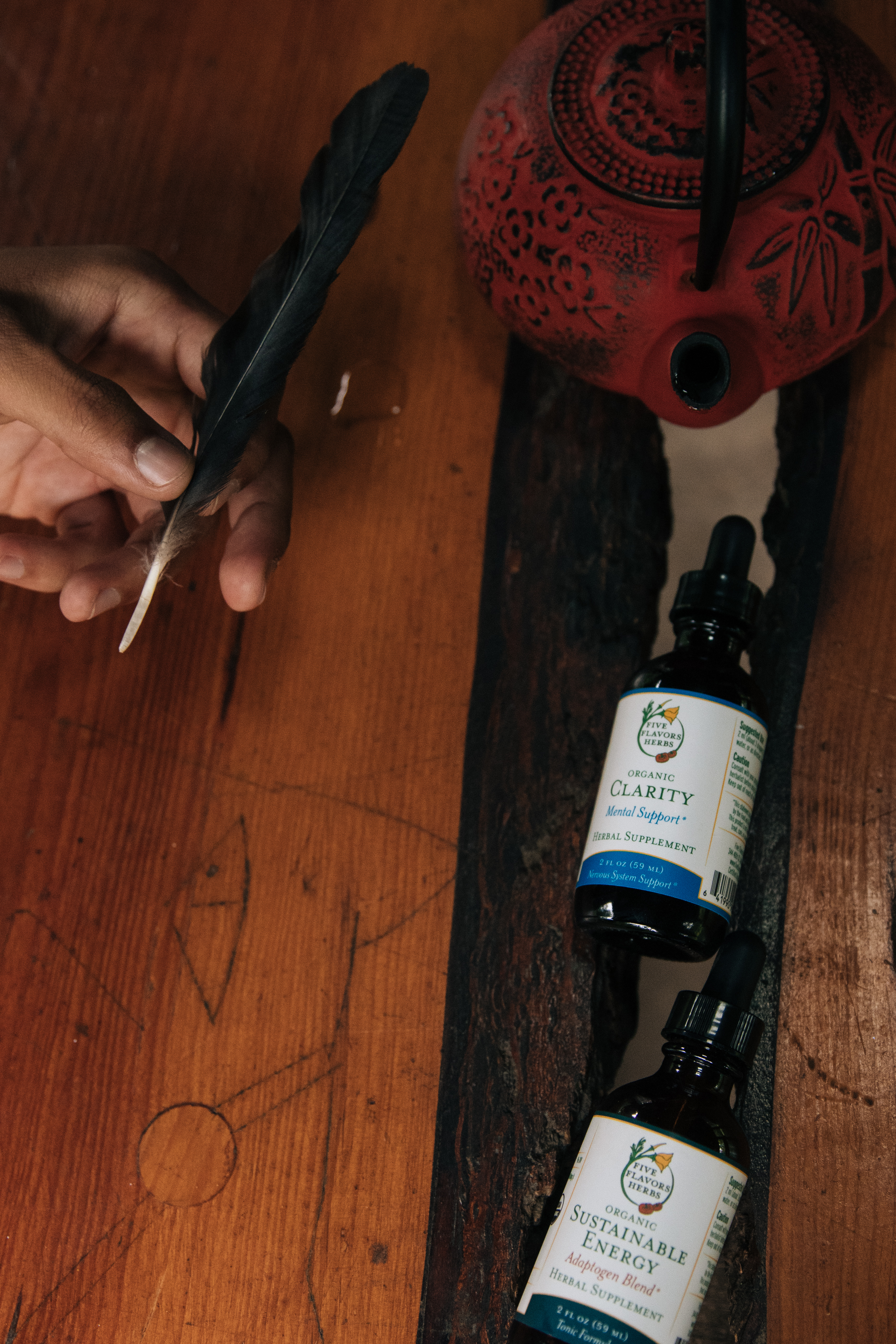hand-holding-black-feather-sitting-on-wooden-table-with-clarity-and-sustainable-energy-tincture-bottles-and-red-and-black-cast-iron-tea-pot-and-laying-next-to-it