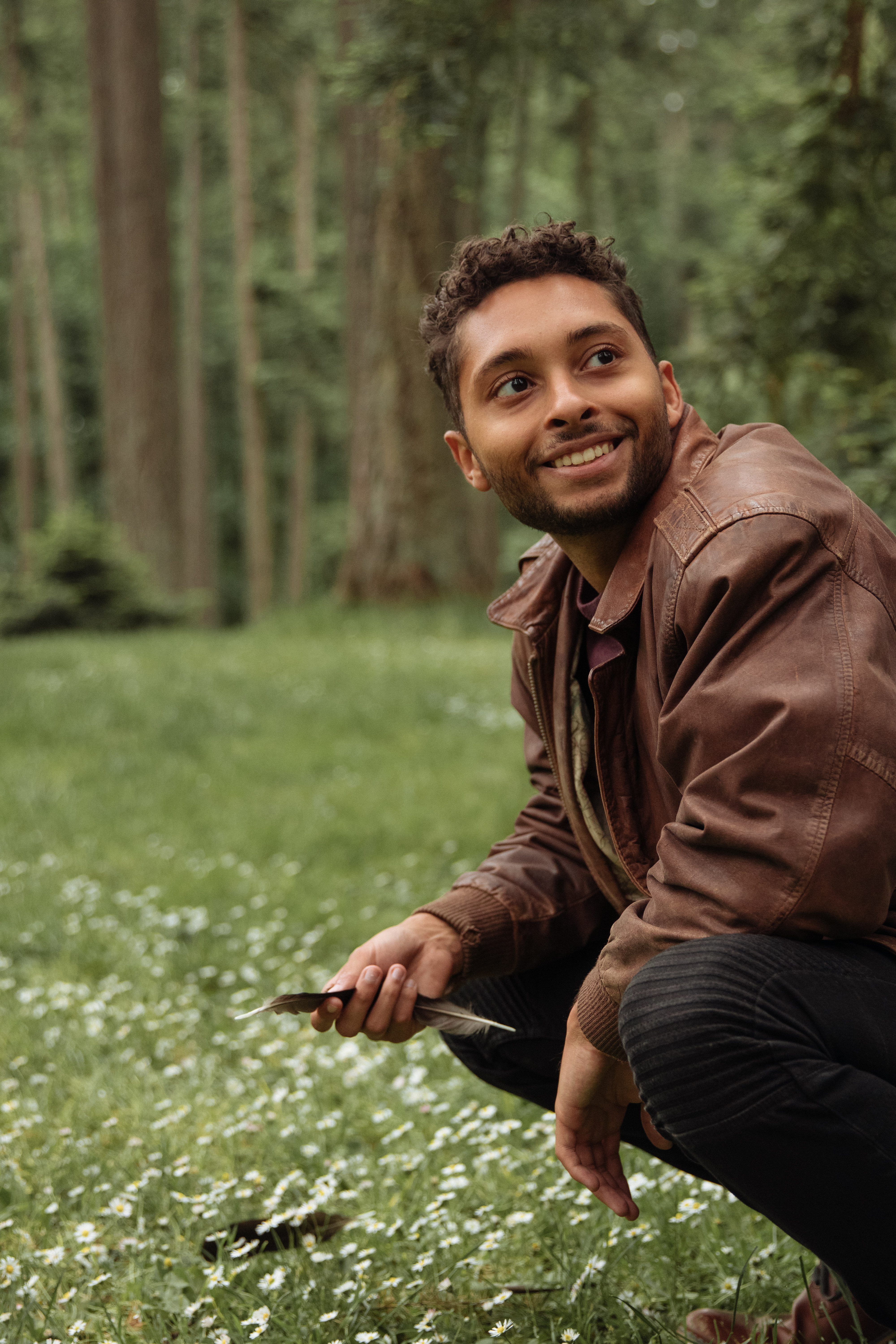 young-man-in-brown-leather-jacket-crouching-in-field-of-wildflowers-with-trees-behind-holding-a-black-feather-and-smiling