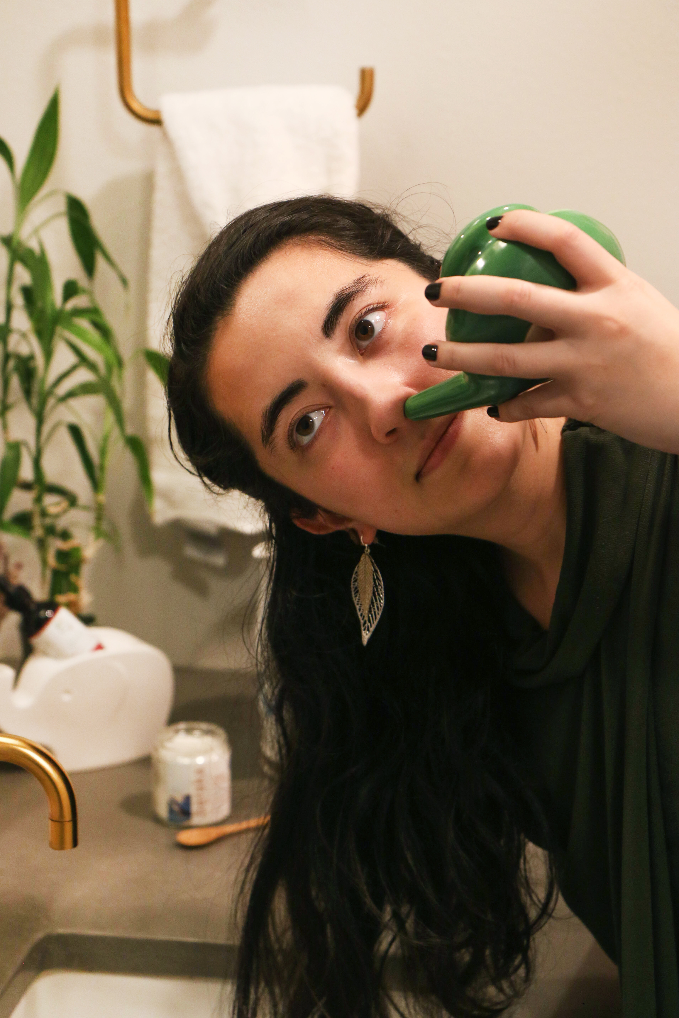 young-woman-with-long-dark-hair-wearing-green-top-and-feather-earrings-leaning-over-bathroom-sink-tipping-green-ceramic-neti-pot-for-sinus-clearing-into-one-nostril