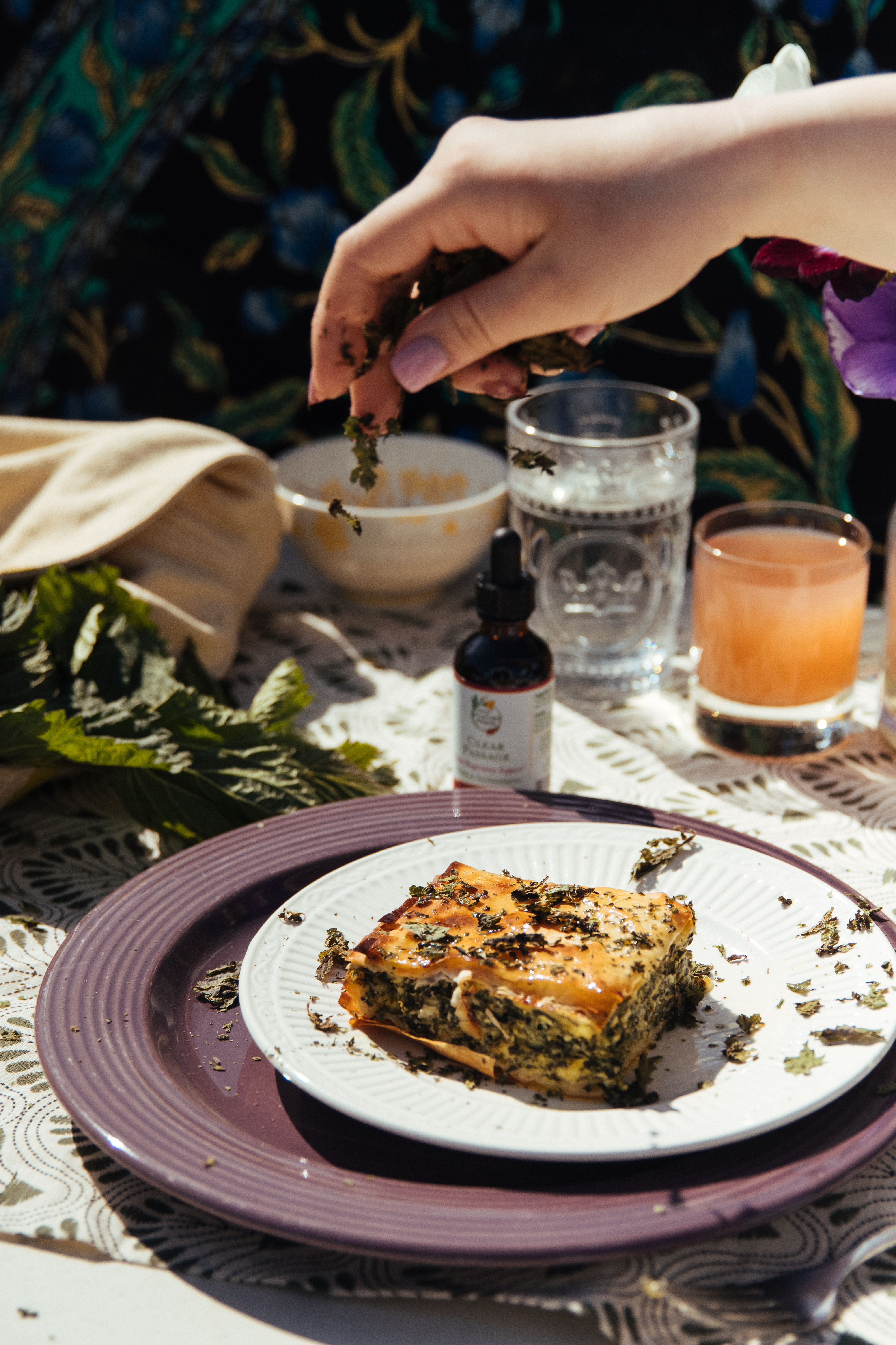 square-piece-of-spanakopita-on-white-plate-on-table-with-drinks-and-fresh-nettles-with-hand-sprinkling-dried-nettle-on-top