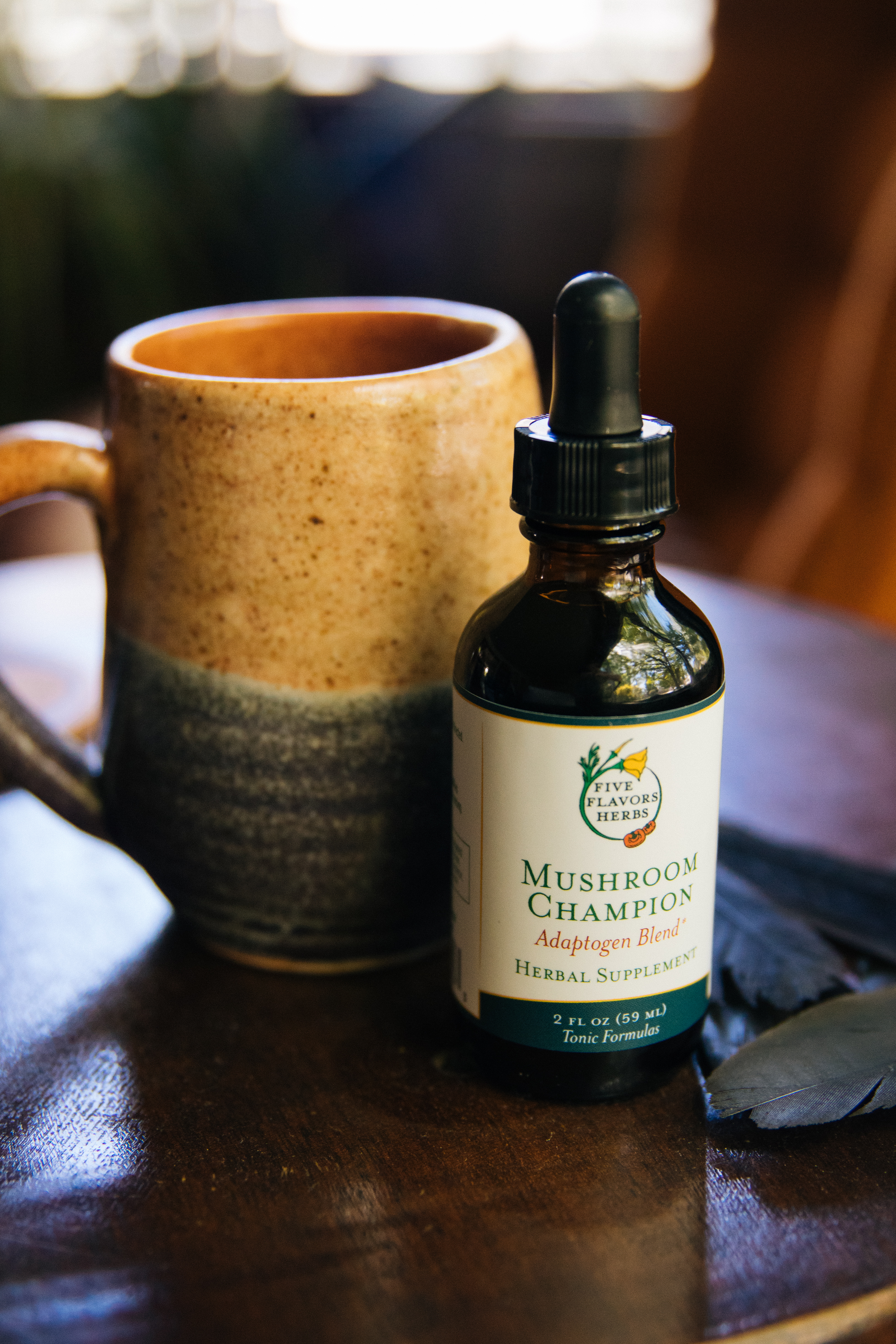 amber-glass-dropper-bottle-of-mushroom-champion-herbal-tincture-sitting-on-wooden-table-next-to-handmade-rustic-pottery-mud-with-living-room-in-background