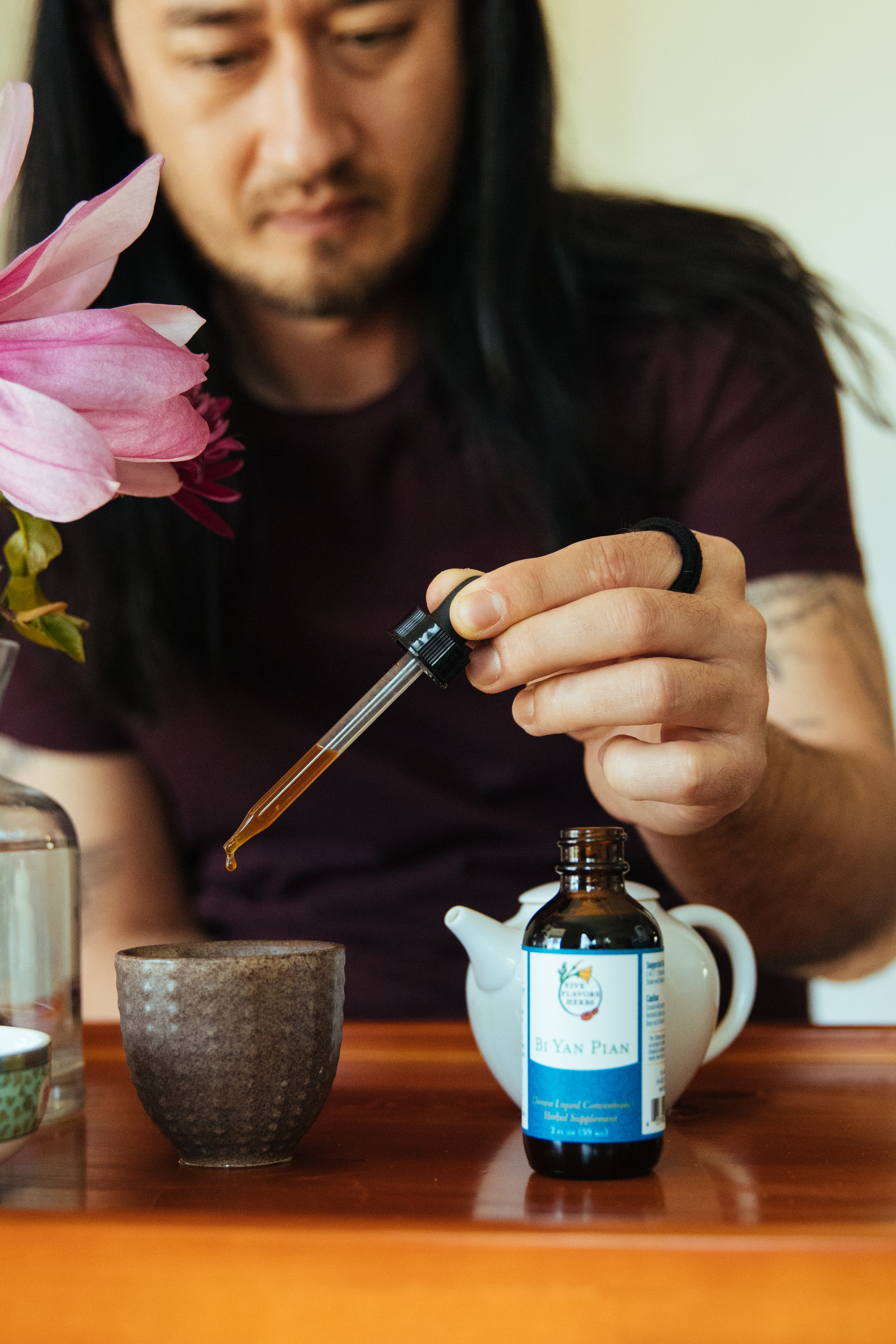 young-man-with-long-hair-dropping-bi-yan-pian-chinese-tincture-formula-for-spring-allergies-into-a-cup-of-tea-with-magnolia-blossoms-to-the-side