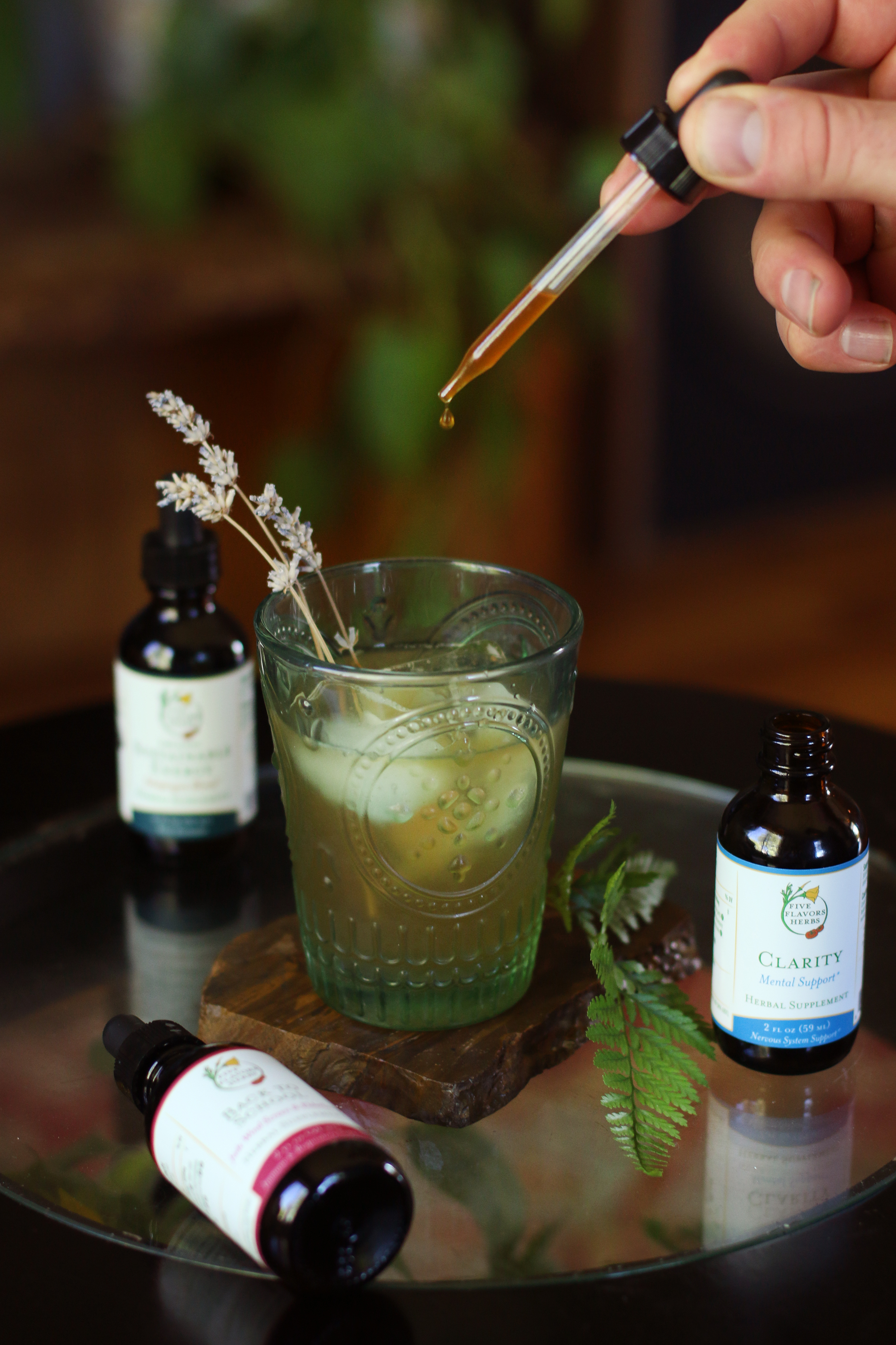 hand-dropping-amber-colored-liquid-tincture-into-vintage-green-glass-surrounded-by-sprigs-of-herbs-and-plants-and-glass-bottles-of-herbal-tincture-blends