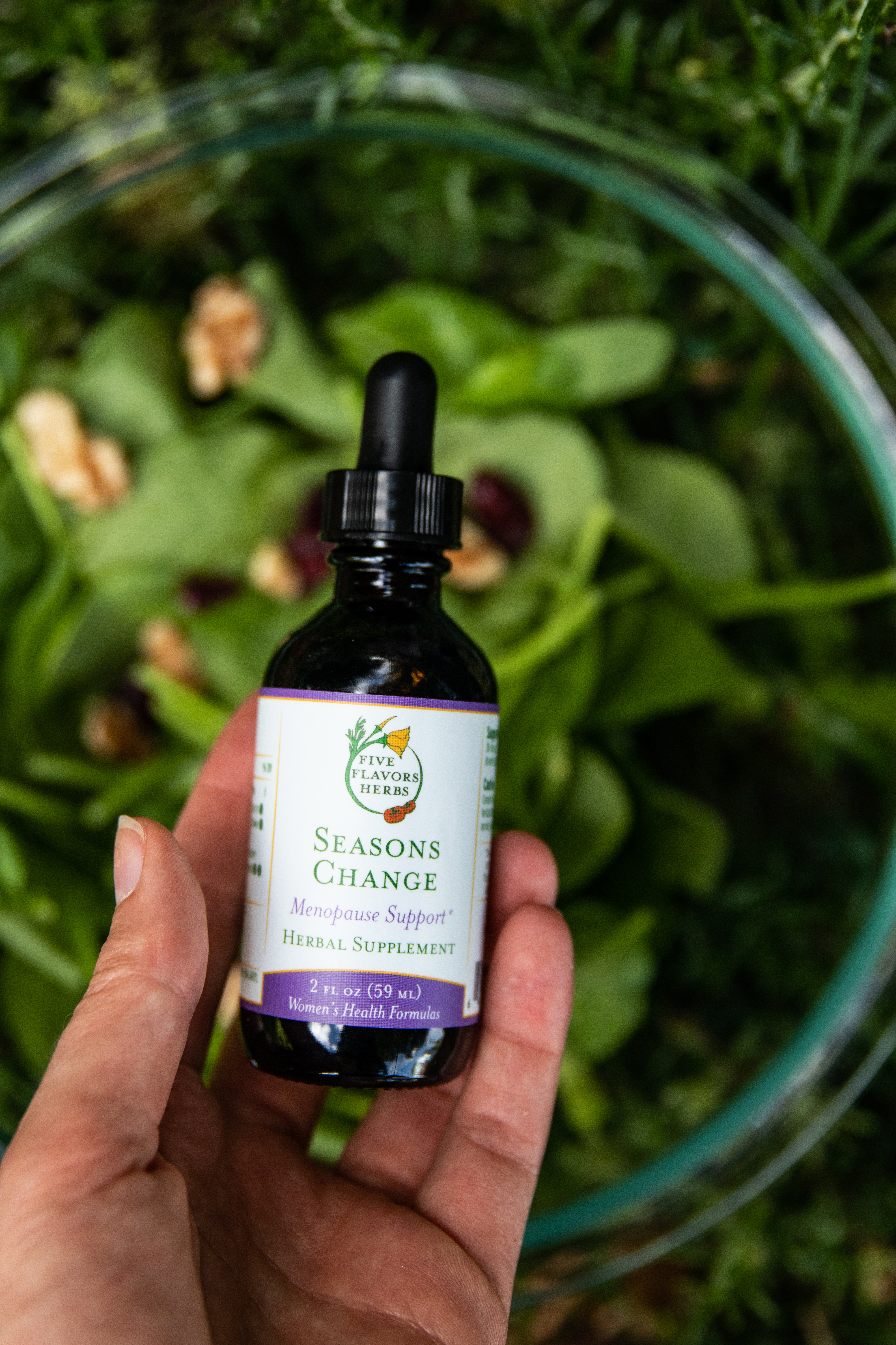 hand-holding-bottle-of-seasons-change-menopause-support-tincture-above-bowl-of-leafy-spinach-greens