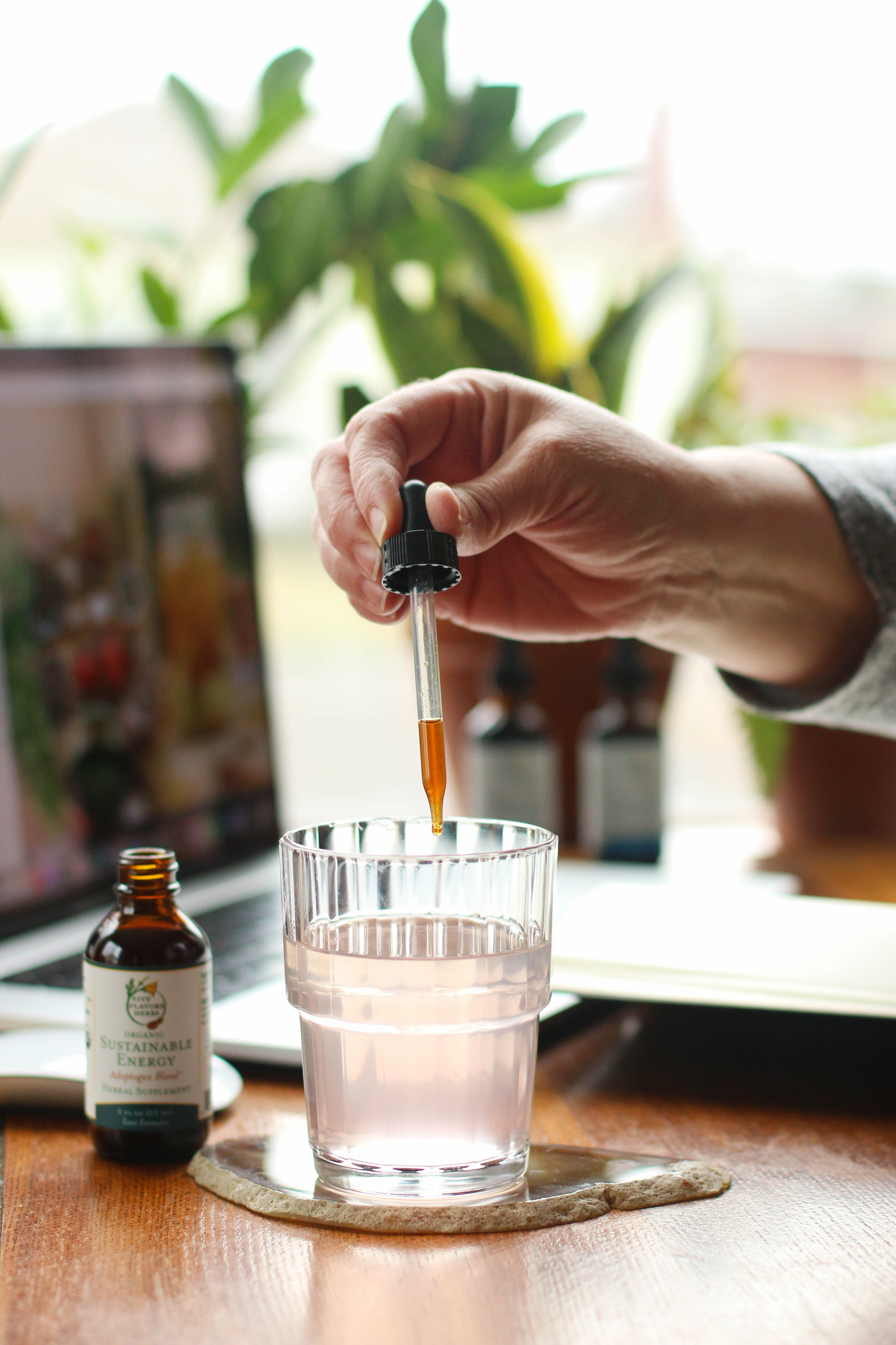 hand-holding-glass-dropper-cap-dropping-tincture-into-glass-of-water-with-work-from-home-desk-setup-in-background